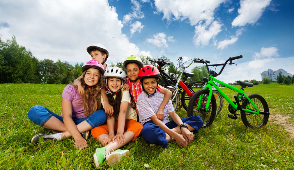 Groupe divers d'adolescents assis dans l'herbe devant leurs vélos, avec leur casque sur la tête | Défi Tête la Première